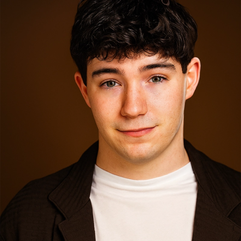 A young man with short, curly dark hair and light skin wears a white shirt and dark jacket. He looks at the camera with a slight, friendly smile. The background is plain brown and softly lit, highlighting Dylan Leiwant’s face and his casual, approachable expression.