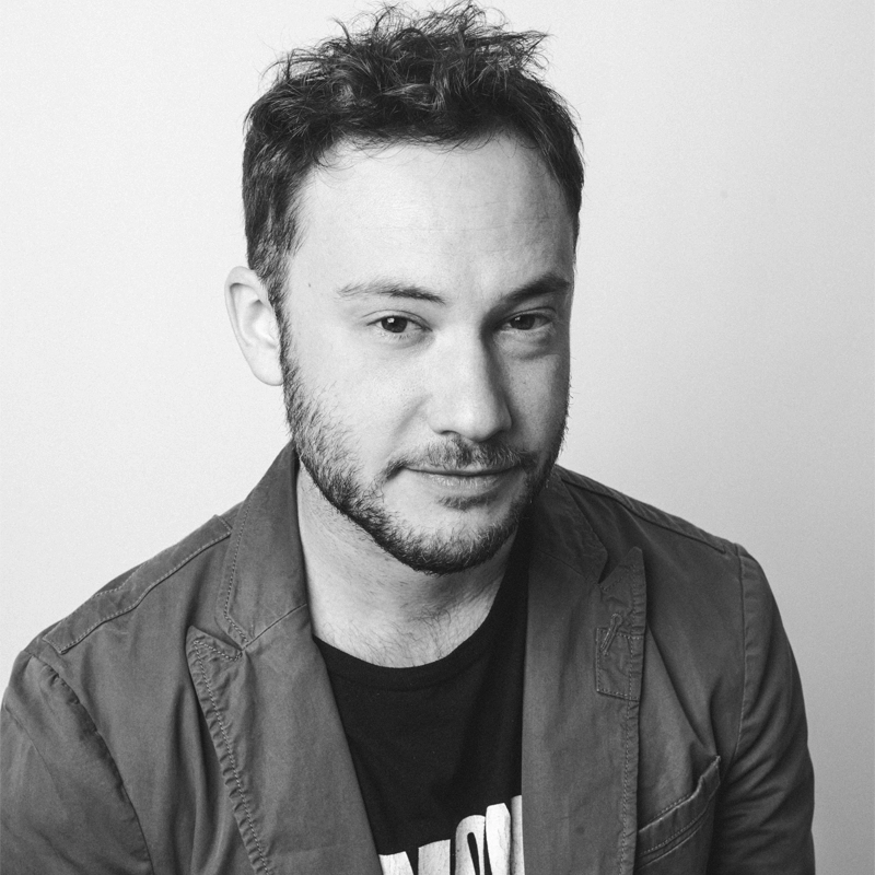 A man with short, tousled hair and a trimmed beard looks at the camera, slightly smiling. He wears a casual jacket over a graphic T-shirt. The photo is black and white, and the background is plain and light-colored.