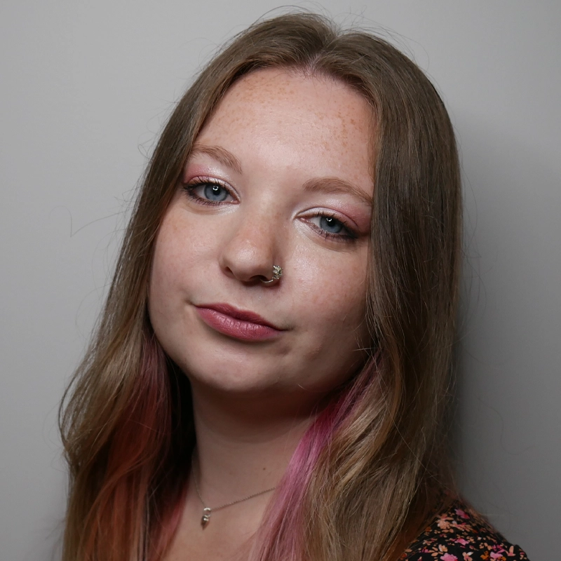 A young woman with light skin and long, straight, light brown hair with pink tips poses in front of a plain light gray background. She has blue eyes, a nose ring, and wears natural makeup, a floral top, and a delicate necklace.