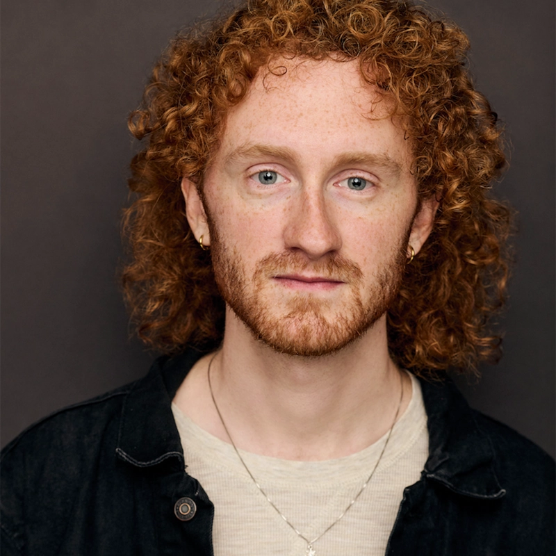 A young man, Harrison Lewis, with curly red hair, light skin, and blue eyes looks at the camera with a neutral expression. He has a short beard, small hoop earrings, a thin necklace, a light shirt, and a black jacket against a plain dark background.