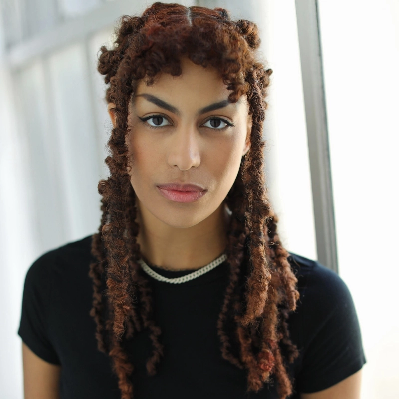A person with medium brown, curly locs styled in twists and space buns stands indoors by a window. Wearing a black top and silver chain necklace, Zoë Lewis-McLean gazes at the camera with a neutral expression as soft natural light illuminates their face.