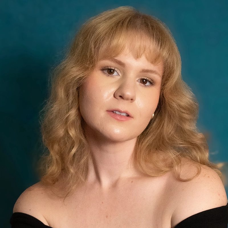 A young person with wavy, shoulder-length blonde hair and bangs poses against a teal background. With fair skin and subtle makeup, Danielle Serrano-Bremer wears an off-the-shoulder black top, gazing softly at the camera with a gentle expression.