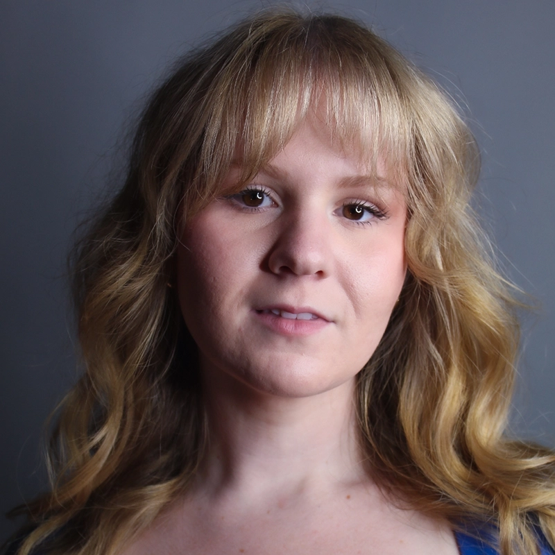A young woman with wavy blonde hair and bangs is standing in front of a plain gray background, looking slightly past the camera with a neutral expression. She is wearing a blue top and has natural makeup. The lighting is soft and even.