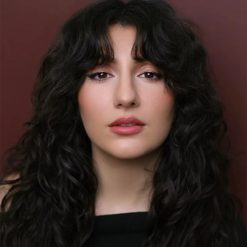 A woman with long, wavy dark hair and bangs poses against a maroon background. She has a light complexion, natural makeup with pink lips, defined eyebrows, and subtle eyeliner. Maggie Likcani looks directly at the camera with a neutral expression.