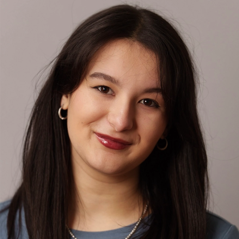 Danielle Serrano-Bremer, a young woman with straight, dark brown hair and fair skin, smiles softly at the camera. She is wearing silver hoop earrings, a blue top, and a silver necklace against a plain, light grey background.