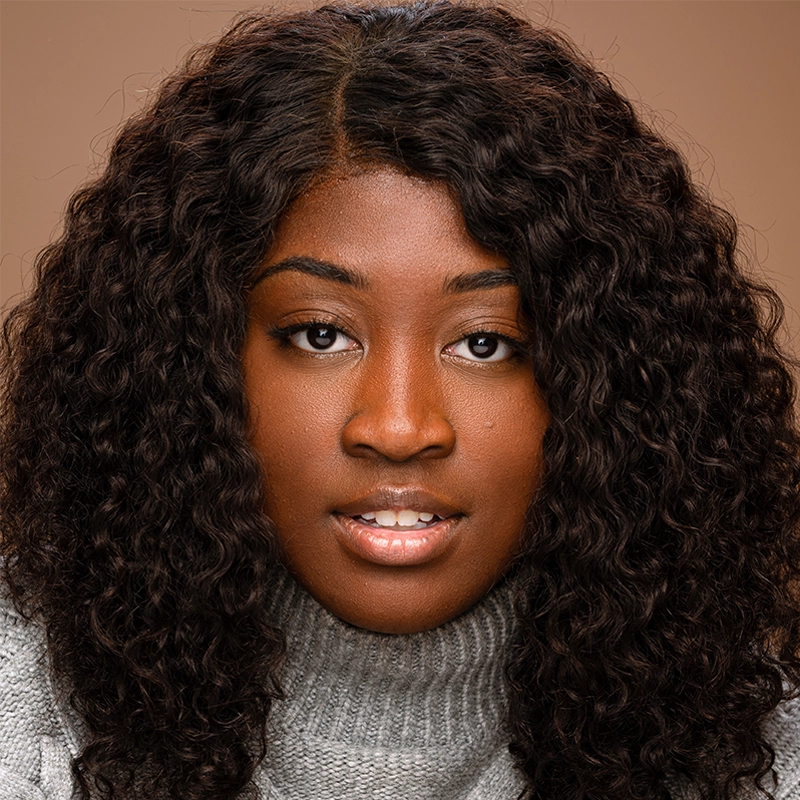 A young woman with deep brown skin and long, curly dark hair looks directly at the camera. She wears a light gray turtleneck sweater. The soft lighting highlights her natural features against a warm brown background, captured by Masa Vujadinovic.