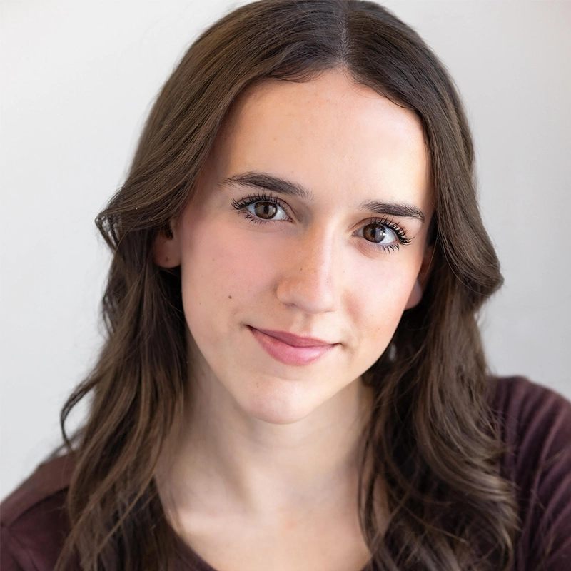 A young woman with long, wavy brown hair and fair skin smiles gently at the camera. She has brown eyes, natural makeup, and wears a dark top. The background is plain and softly lit, highlighting her face.