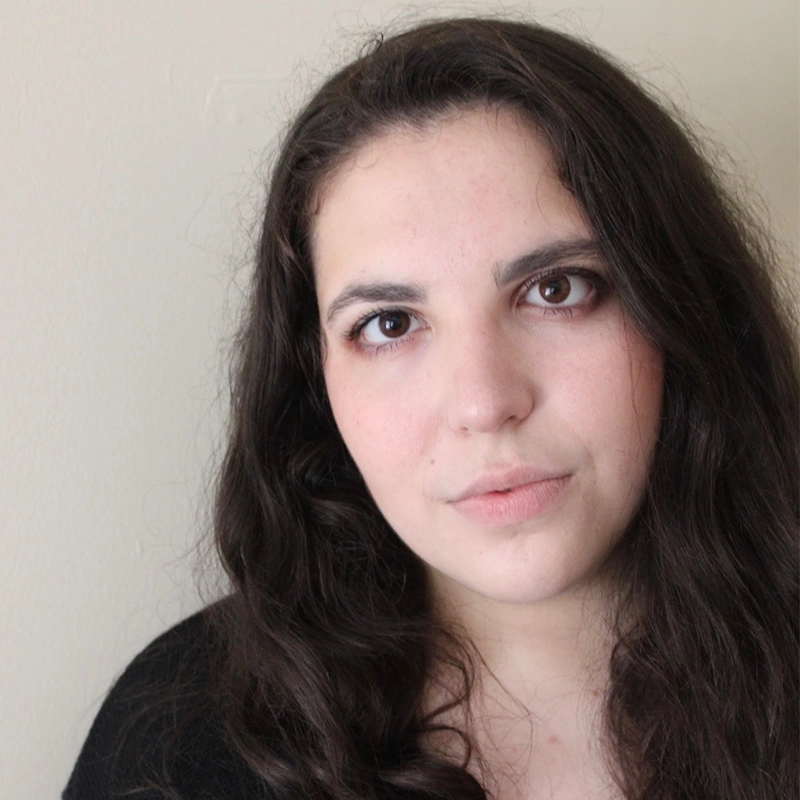 Caroline Weston, with long, wavy dark hair and fair skin, looks directly at the camera with a neutral expression. She is wearing a black top and minimal makeup against a plain, light-colored background.