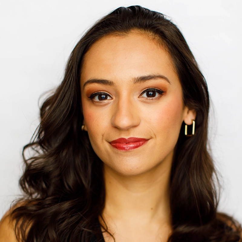 A woman with wavy dark brown hair and light brown skin smiles gently. She wears gold hoop earrings, red lipstick, and warm-toned eye makeup. Posed against a plain white background, she exudes RJ Vandenbrouck’s signature elegance and looks directly at the camera.
