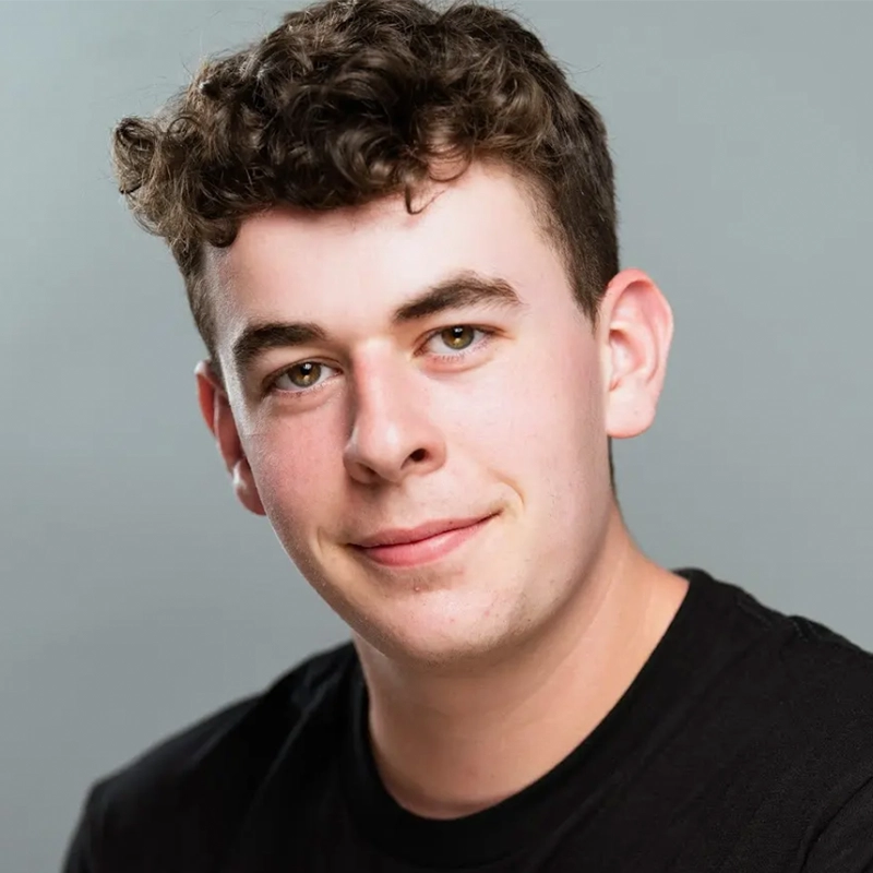 A young man with short, curly brown hair and fair skin is smiling softly at the camera. He is wearing a plain black t-shirt and is posed against a simple, light gray background. The lighting is even and natural.