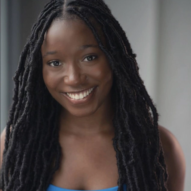 Kayla Louison, a young woman with long, dark, twisted locks, smiles warmly at the camera. She wears a blue top and stands indoors against a softly blurred gray background, creating a natural and inviting portrait.