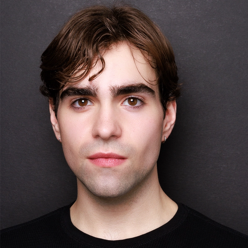 A young man with fair skin, brown eyes, and short brown hair poses against a dark background. Rynn Stephenson wears a black shirt and a small hoop earring in his left ear, gazing directly at the camera with a neutral expression.