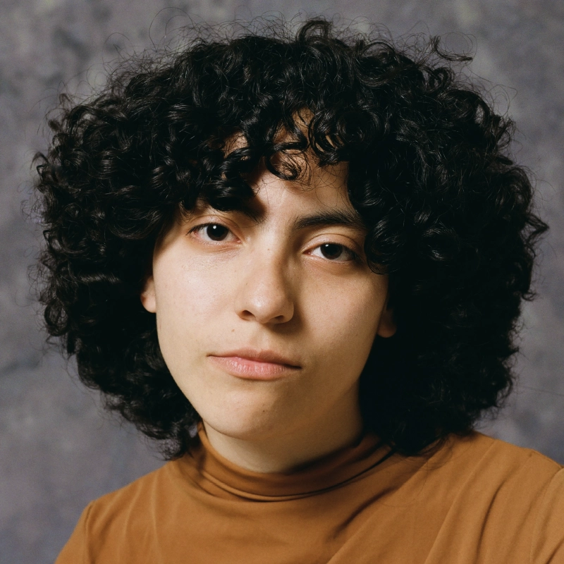 A person with medium skin, dark curly hair, and brown eyes looks at the camera with a neutral expression. Danielle Serrano-Bremer is wearing a light brown turtleneck. The background is a soft, mottled gray.