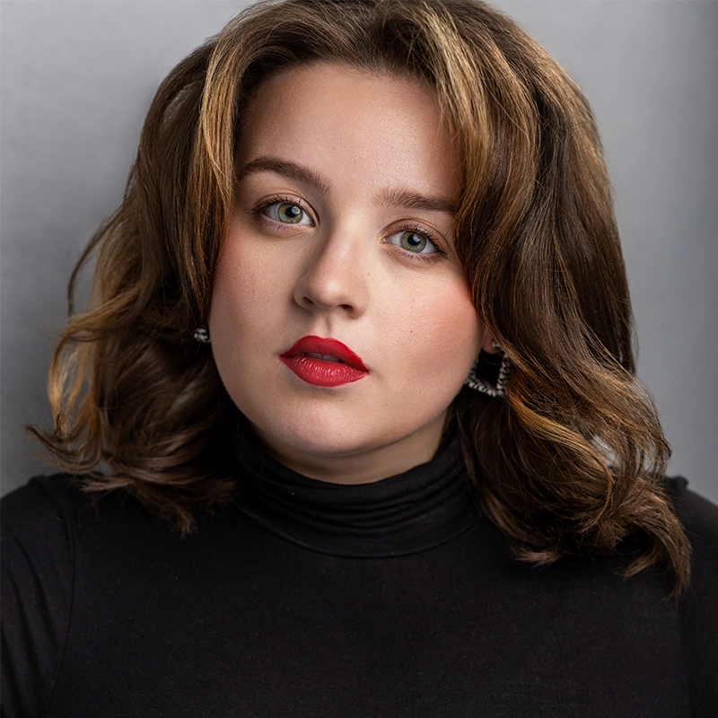 Sahara Williams, a young woman with wavy brown hair and highlights, wears a black turtleneck top. She has fair skin, bold red lipstick, subtle makeup, and sparkly square earrings, looking directly at the camera against a gray background.