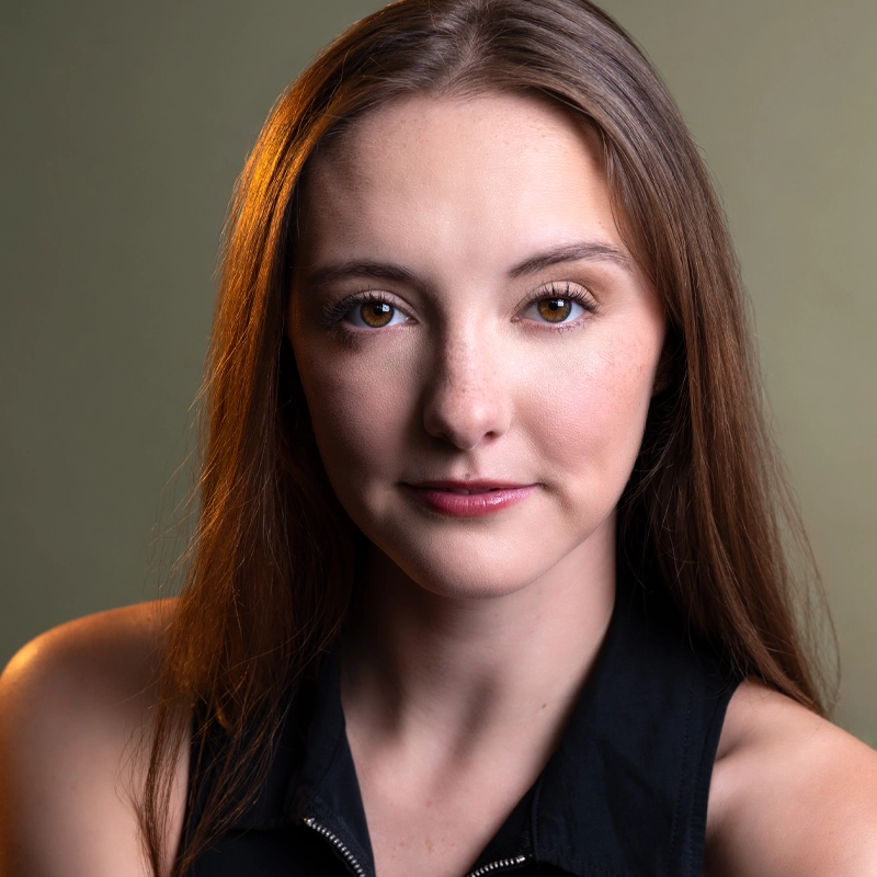 Kelsey MacDonald, a young woman with long brown hair and light skin, looks directly at the camera, softly smiling. She wears a sleeveless black top. The muted green background and warm light highlight the left side of her face and hair.
