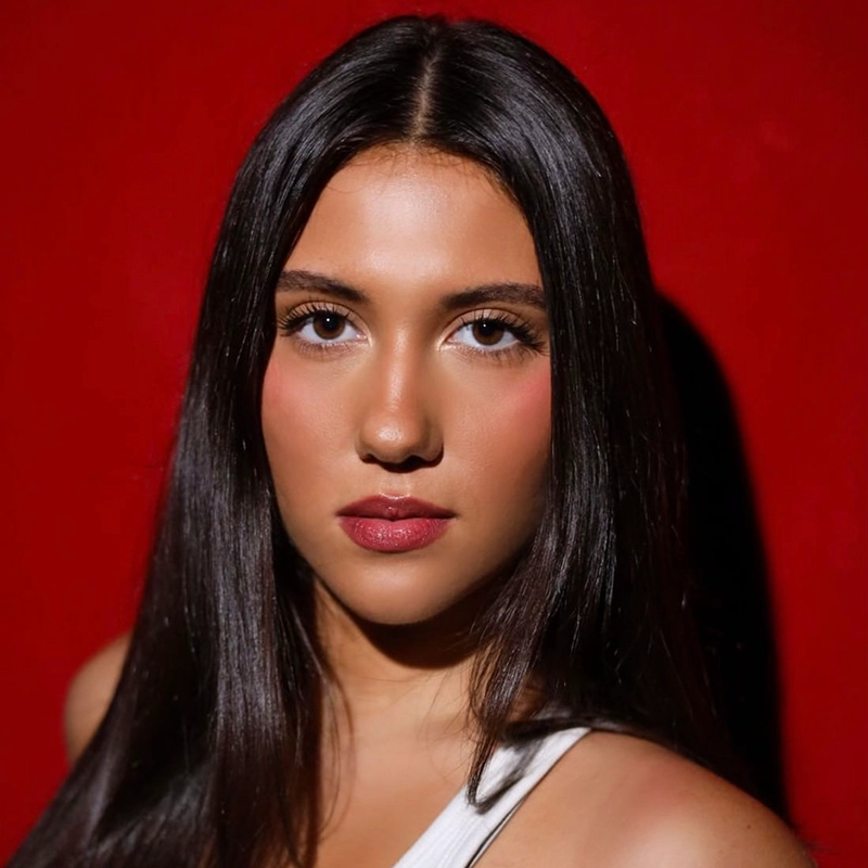 Madelyn Martinez, with long, straight dark hair and glowing skin, looks directly at the camera. She wears a white sleeveless top and natural makeup with rosy cheeks and lips. The deep red background creates a striking contrast with her complexion.