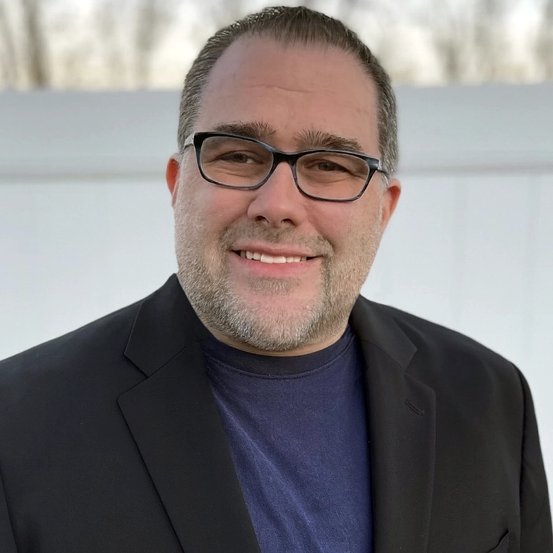 Joe Marchese, a middle-aged man with short brown hair, glasses, and a trimmed beard, smiles at the camera. He is wearing a dark blazer over a navy blue shirt, set against a softly blurred outdoor background with a white fence.