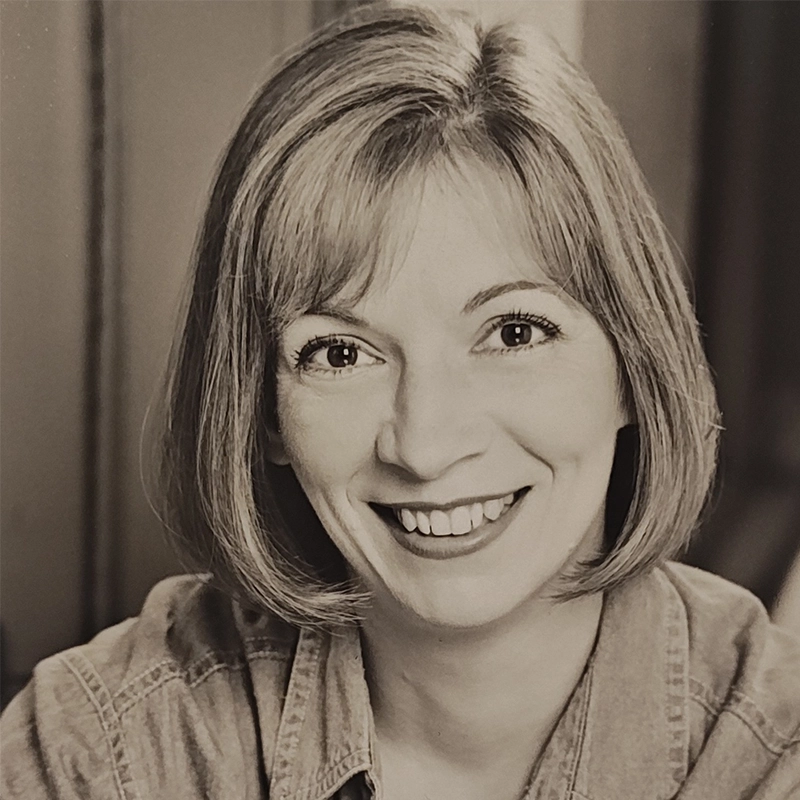 A black-and-white portrait of Caroline Weston, a smiling woman with straight, chin-length hair and side-swept bangs. She wears a denim shirt and faces the camera directly, exuding warmth and friendliness against a softly blurred background.
