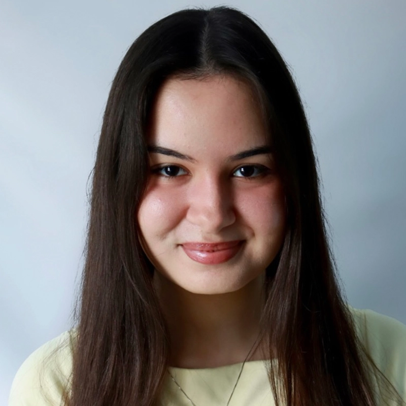A young woman with long straight dark hair, light skin, and brown eyes smiles gently at the camera. Danielle Serrano-Bremer wears a cream-colored top and a thin necklace, set against a plain, light background for a clean, professional look.
