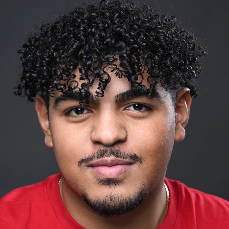 Young man, Dayven Martinez, with curly black hair, trimmed beard, and mustache smiles slightly. He wears a red shirt and thin gold chain. The dark background highlights his smooth skin and expressive eyes under soft lighting.