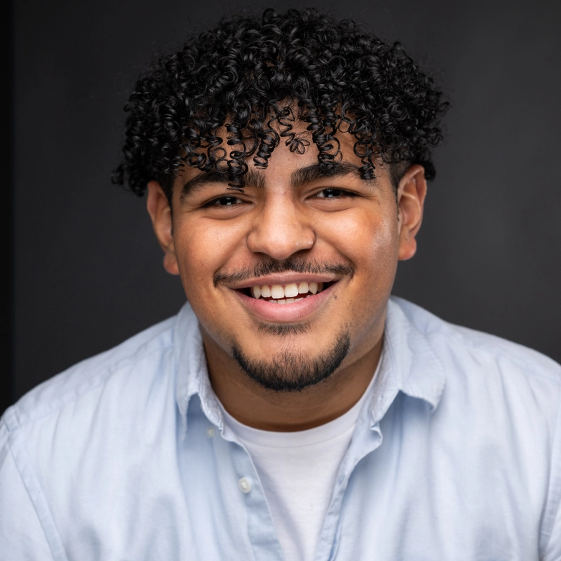 Dayven Martinez, a young man with curly black hair, a trimmed goatee, and a mustache, smiles warmly at the camera. He wears a light blue button-down over a white t-shirt. The dark background highlights his cheerful expression.