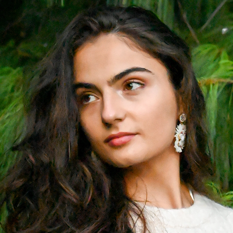 A young woman with long brown wavy hair looks thoughtfully to her left. She wears a white top and large decorative earrings. Green foliage fills the blurred background, creating a serene, natural atmosphere—an elegant portrait by Masa Vujadinovic.