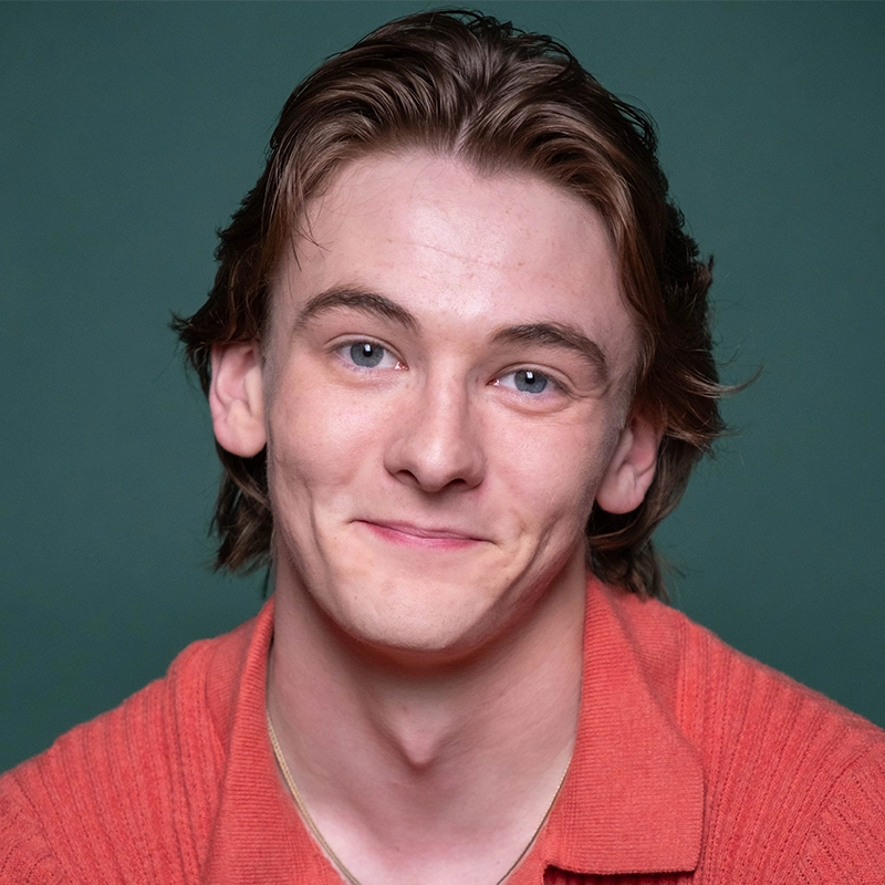 A young man with fair skin, blue eyes, and medium-length light brown hair smiles softly. He is wearing a textured coral-orange shirt against a plain dark green background. His head is slightly tilted, and he has visible dimples on his cheeks.