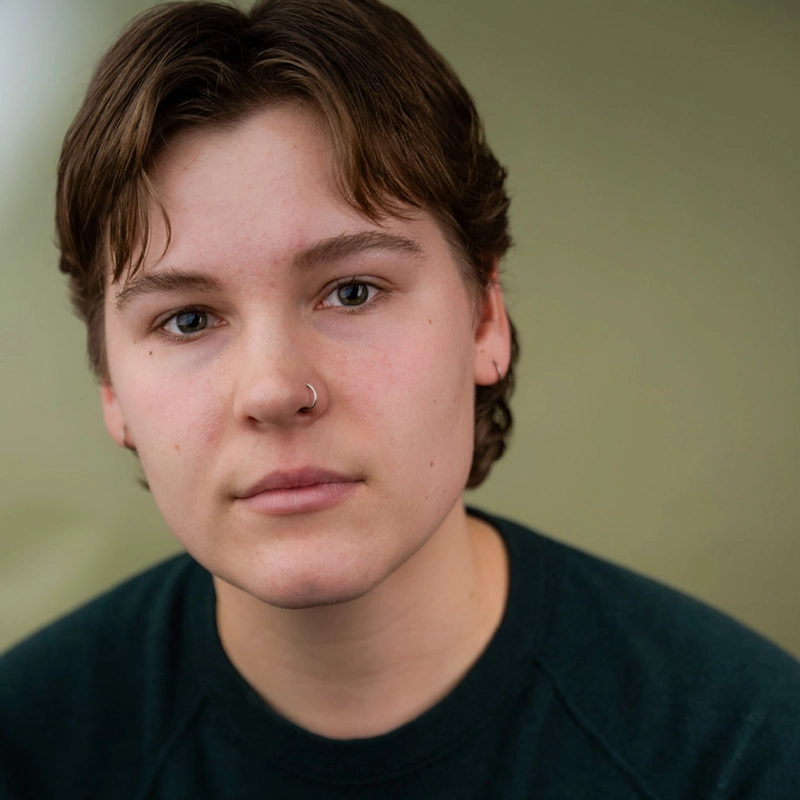 A person with short brown hair, light skin, and a nose ring gazes softly at the camera. They wear a dark green sweater and are posed against a plain, muted green background. Their expression is calm and neutral.