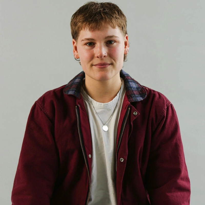 A person with short brown hair wears a burgundy jacket with a plaid collar over a white T-shirt. They have small hoop earrings and a silver necklace. They are smiling gently and sitting against a plain light gray background.