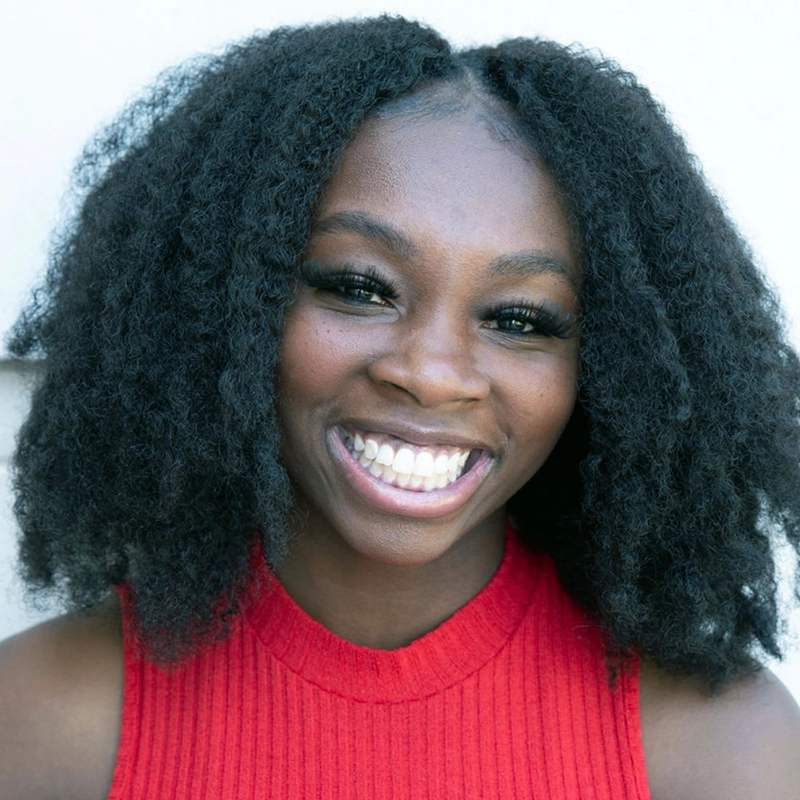 Lauren Torey smiles brightly, showing her teeth. A young woman with natural curly black hair, she wears a sleeveless, ribbed red top and has dramatic eyelashes. The background is a plain, light-colored wall.