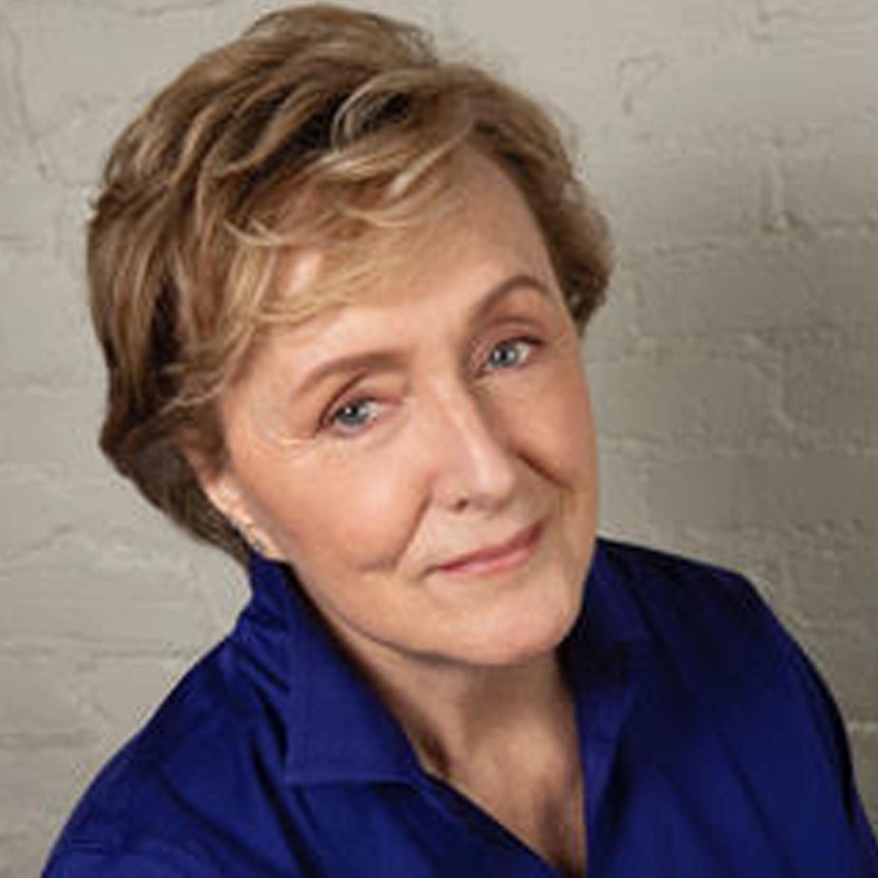 Annie McGreevey, with short, wavy light brown hair, wears a deep blue collared shirt and smiles gently. The off-white, textured brick wall background and soft lighting create a warm, approachable appearance.