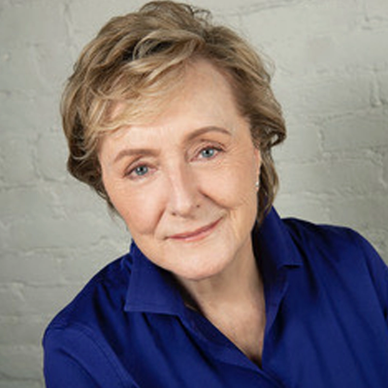 Annie McGreevey, an older woman with short, light brown hair and blue eyes, smiles gently. She wears a royal blue collared shirt and small earrings. The softly lit background features a light gray, textured brick wall.