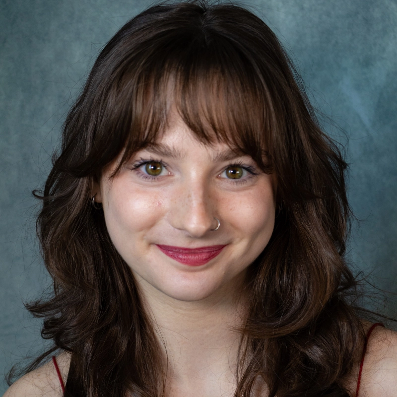 A young woman, Ragan McKenna, with wavy brown hair and bangs smiles softly. She has light skin, hazel eyes, a nose ring, and wears subtle makeup with red lipstick. The background is a soft blue-gray gradient.