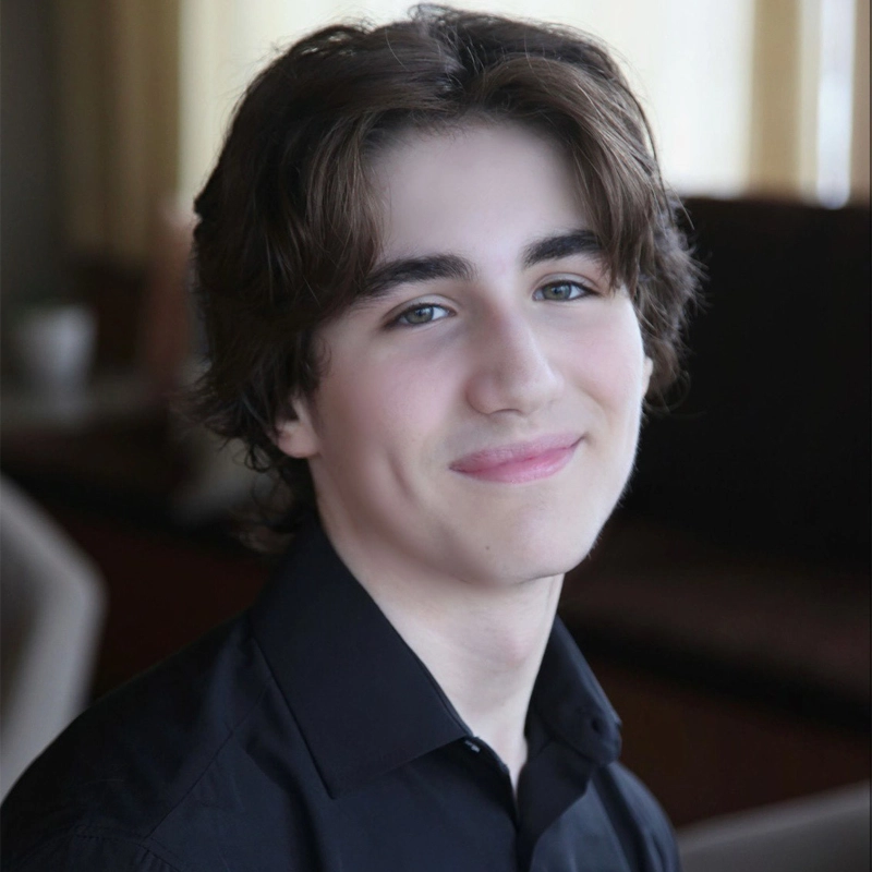 A young person with medium-length brown hair and fair skin smiles softly at the camera. Harrison McNeill wears a black collared shirt and sits in a softly lit indoor setting with blurred furniture and cream-colored curtains in the background.