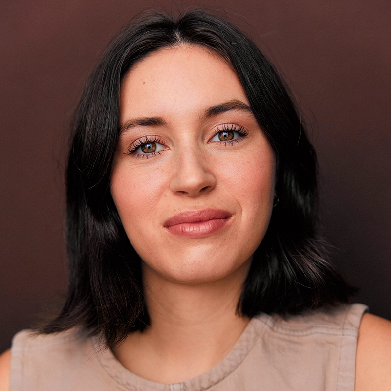 A woman with shoulder-length dark hair and light skin, resembling Mickey Wirtz, looks at the camera, smiling softly. She wears a sleeveless beige top and natural makeup, with a brown background behind her. Her expression is calm and confident.