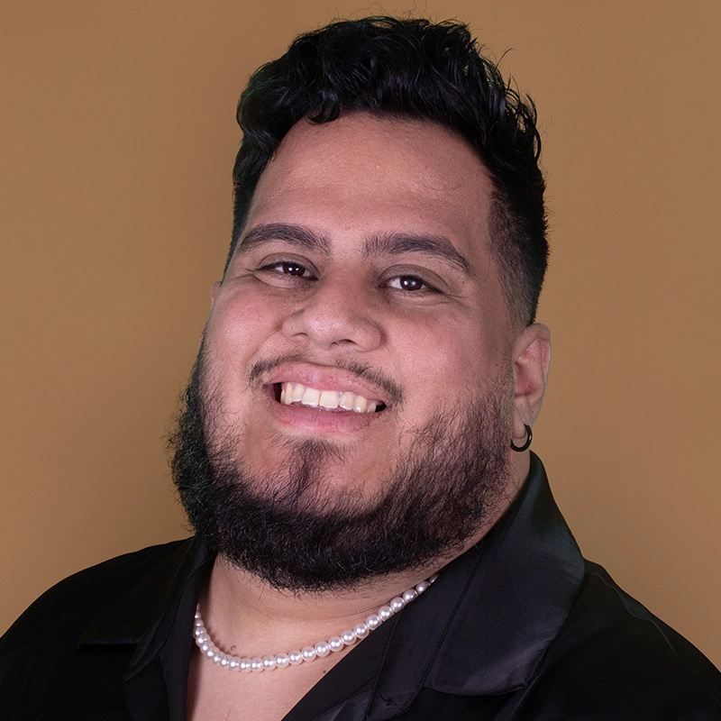 A smiling Danielle Serrano-Bremer with short, dark, wavy hair and a full beard wears a black shirt, pearl necklace, and small hoop earring. The solid light brown background highlights their confidence and warmth as they face the camera.