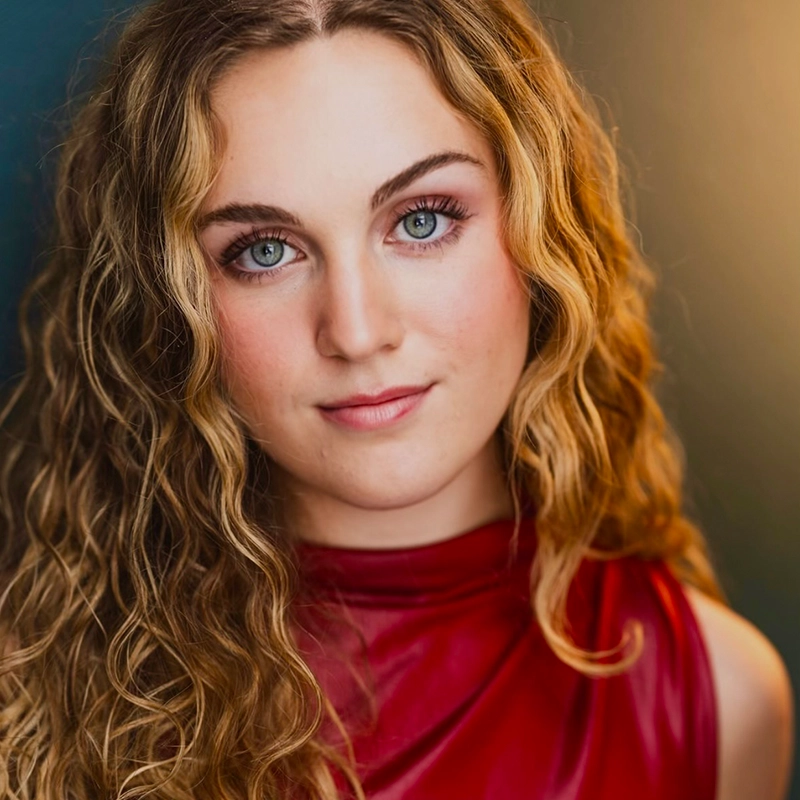 A young woman with long, wavy blonde hair and blue eyes looks at the camera with a slight smile. Wearing a sleeveless, shiny red top, she channels Mickey Wirtz’s effortless style against a softly blurred backdrop of warm and cool tones.
