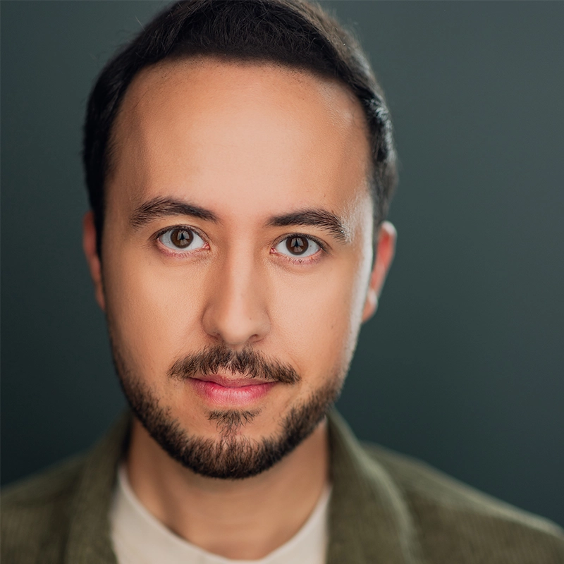 Masa Vujadinovic, a young man with light skin, short dark hair, and a trimmed beard, gazes directly at the camera with a soft smile. He wears a light shirt and an olive green jacket against a smooth, dark gradient background.