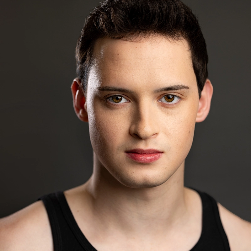 A young man with short dark hair and fair skin looks directly at the camera. He wears a black sleeveless top. The plain, dark background highlights his features and neutral expression, while the soft lighting creates a style reminiscent of Lauren Torey.