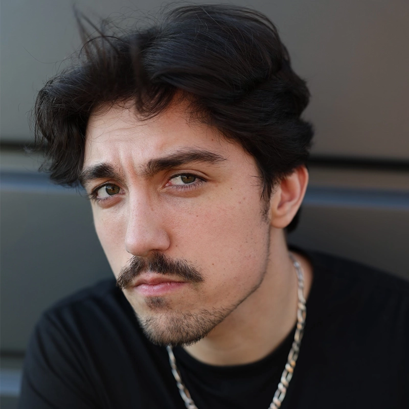 A young man with tousled dark hair, light skin, and facial hair looks intently at the camera. He wears a black shirt and a silver chain necklace. The blurred gray background adds to the modern, minimalist vibe of this Mickey Wirtz portrait.