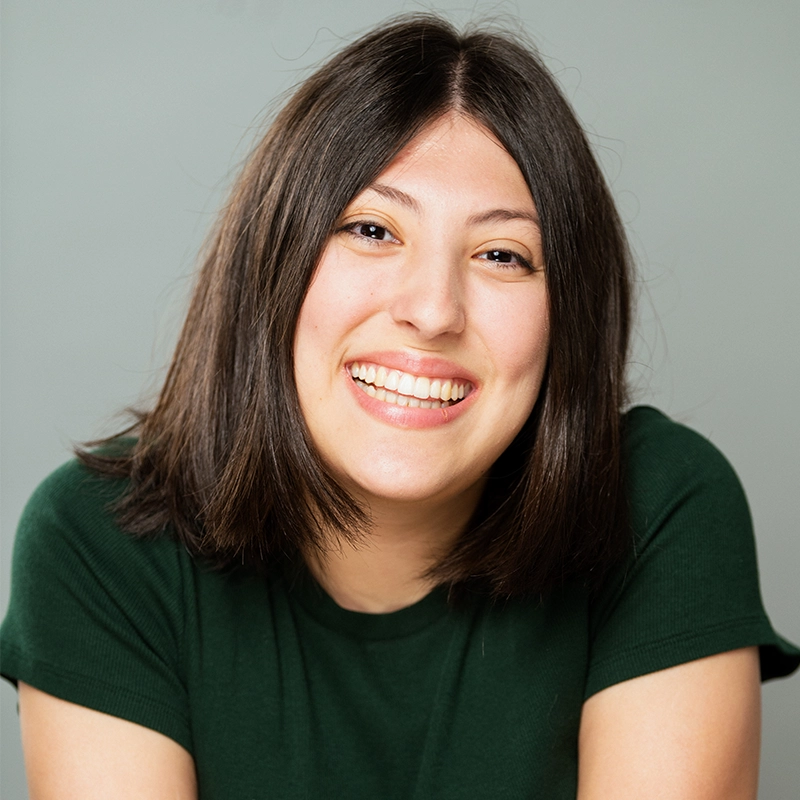 Sahara Williams, a young woman with straight, shoulder-length brown hair, smiles broadly at the camera. She wears a dark green short-sleeved shirt and sits before a plain, light gray background, her expression cheerful and friendly.