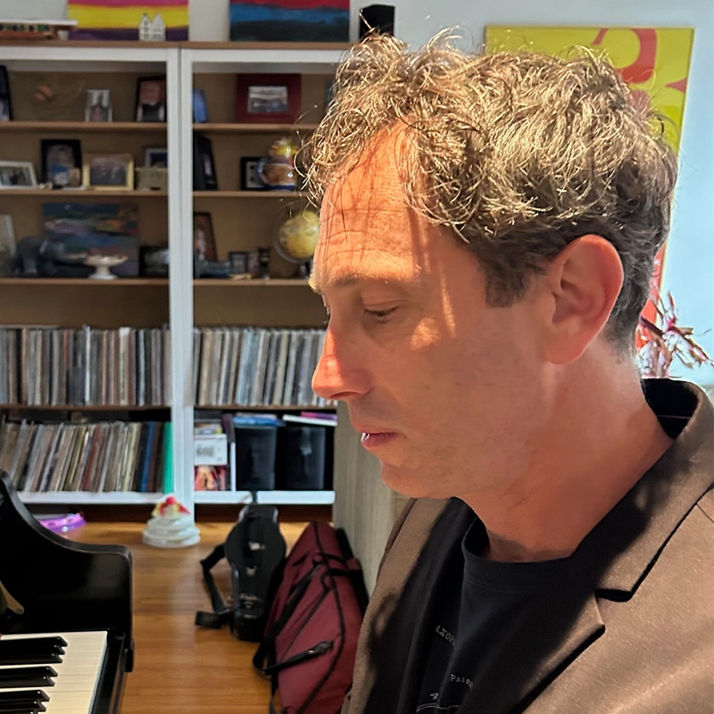A man with curly hair wearing a dark blazer looks down beside a piano. Behind him, shelves hold records, books, globes, and framed photos—some by Hannah Thompson. The warmly lit scene exudes a cozy, creative atmosphere filled with personal touches.