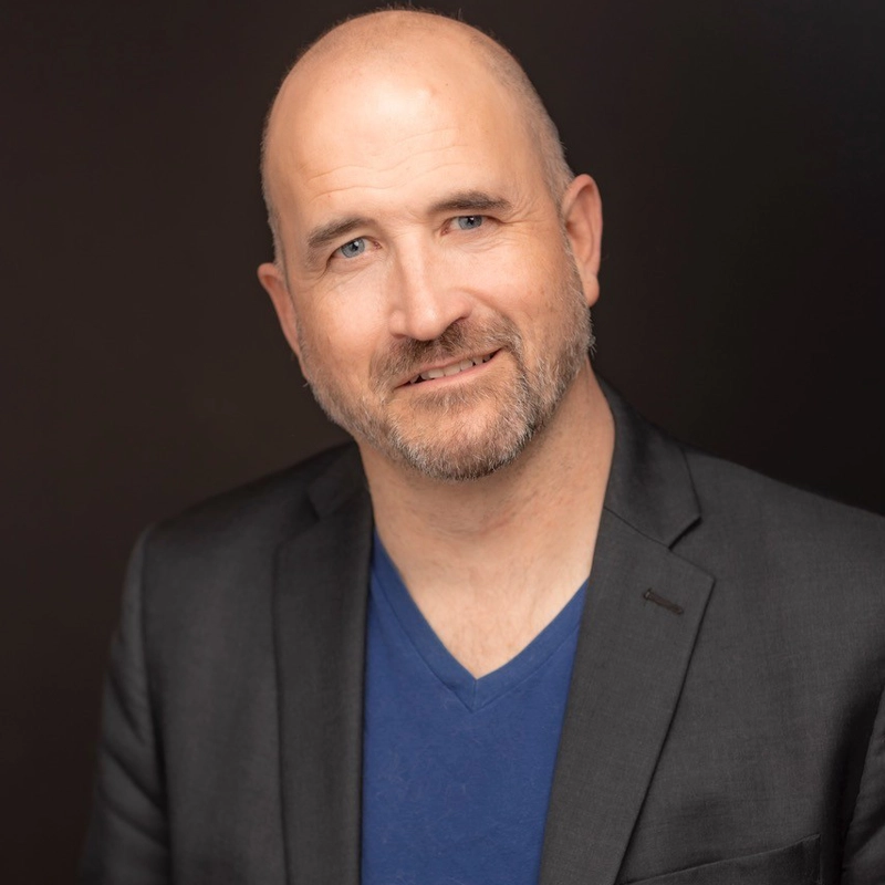 Travis Mitchell, a middle-aged man with a bald head and trimmed beard, smiles slightly. He wears a black blazer over a blue V-neck shirt. The dark, plain background highlights his friendly, approachable expression.