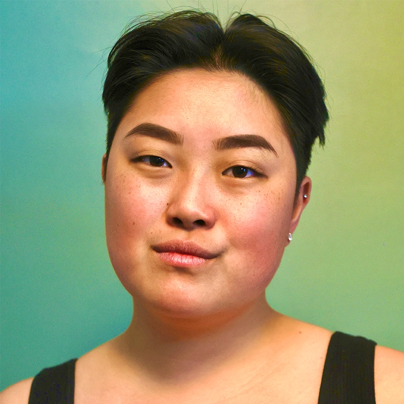 A person with short dark hair and smooth skin looks confidently at the camera. They wear a black tank top and small earrings. The expression is neutral with slightly pursed lips. The background fades from turquoise on the left to yellow-green on the right.