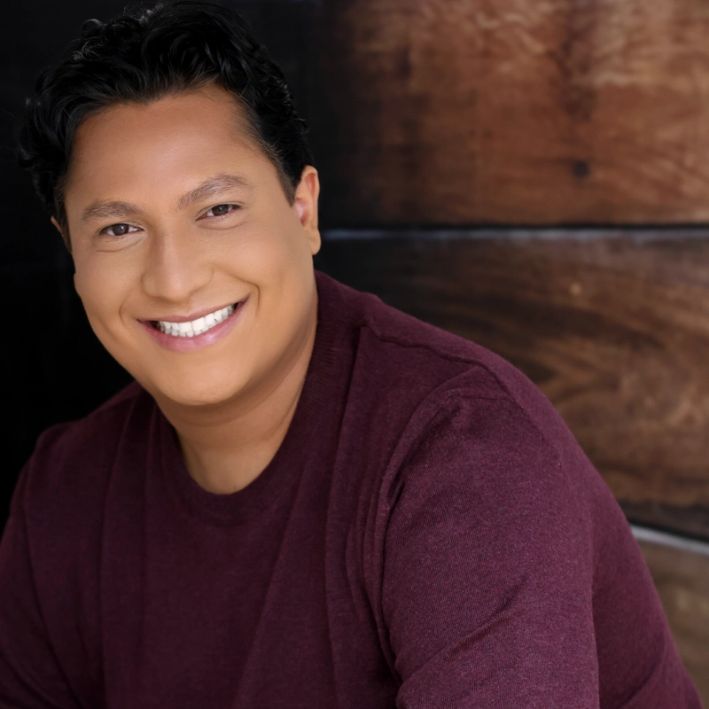 Kyle Morales, with short dark hair and medium skin tone, smiles brightly at the camera. Wearing a burgundy shirt, Kyle is posed before a warm wooden background, and the soft lighting creates a welcoming and friendly atmosphere.