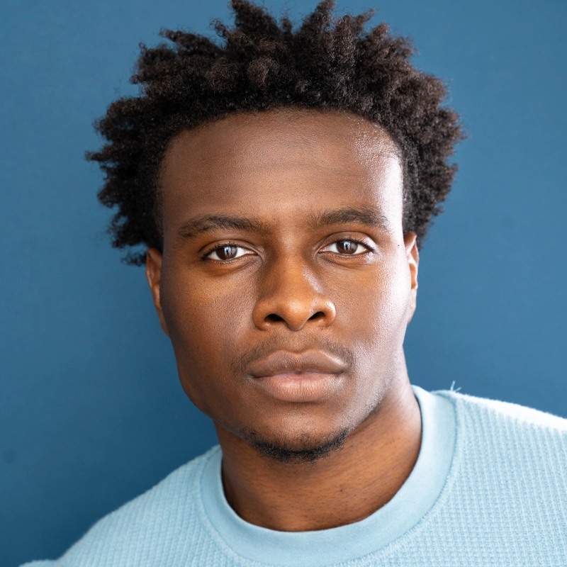 A young Black man with short, curly hair stares directly at the camera with a serious expression. He is wearing a light blue textured sweater and is posed against a solid blue background. The lighting is soft and even, highlighting his facial features.