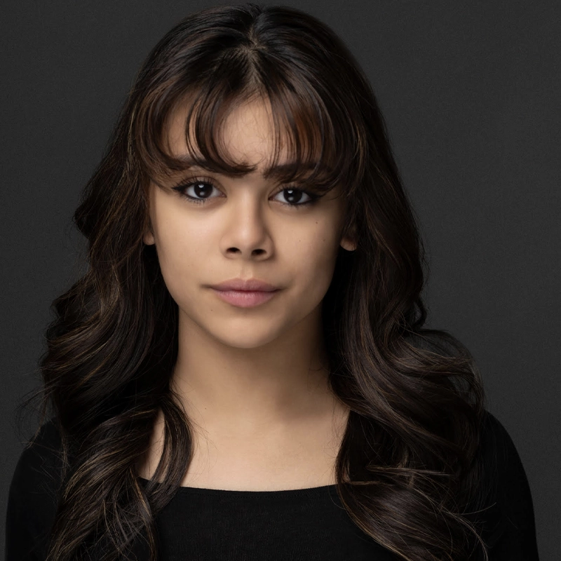 A young woman with long, wavy dark brown hair and bangs poses against a dark gray background. She has a neutral expression, with natural makeup and dark eyes. She is wearing a simple black top. The lighting is soft and even.