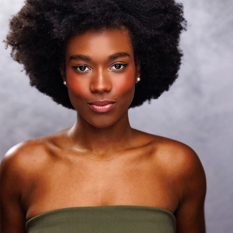 Caroline Bowers, with natural afro hair and glowing skin, gazes confidently at the camera. She wears subtle makeup with blush and nude lipstick, pearl earrings, and an olive green, off-shoulder top against a soft, cloudy gray background.
