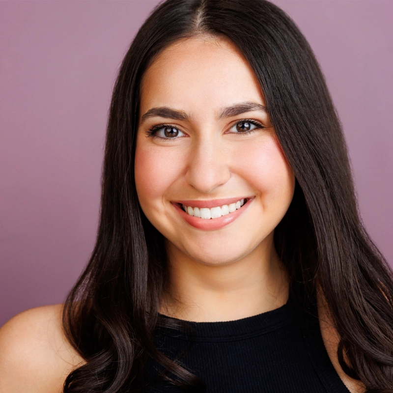 Danielle Nodelman, a young woman with long, dark brown hair, smiles warmly at the camera. She has smooth, light skin and brown eyes and wears a sleeveless black top against a solid mauve background for a professional, friendly look.