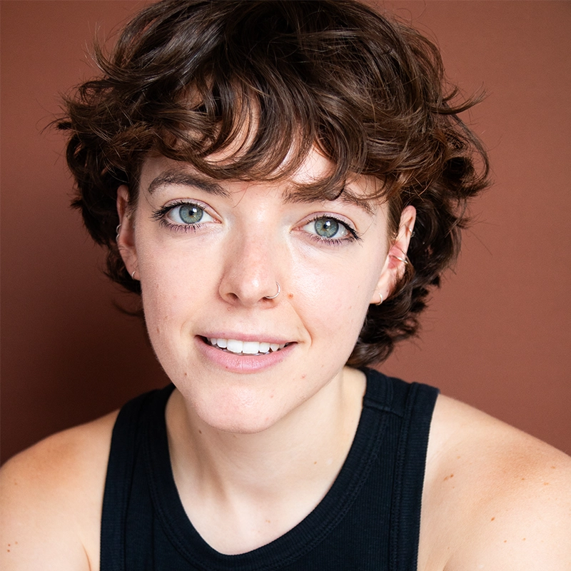 A young person with short, curly brown hair and light skin smiles softly at the camera. Styled by Caroline Bowers, they wear a black sleeveless top against a plain brown background, giving the image a warm and natural feel.